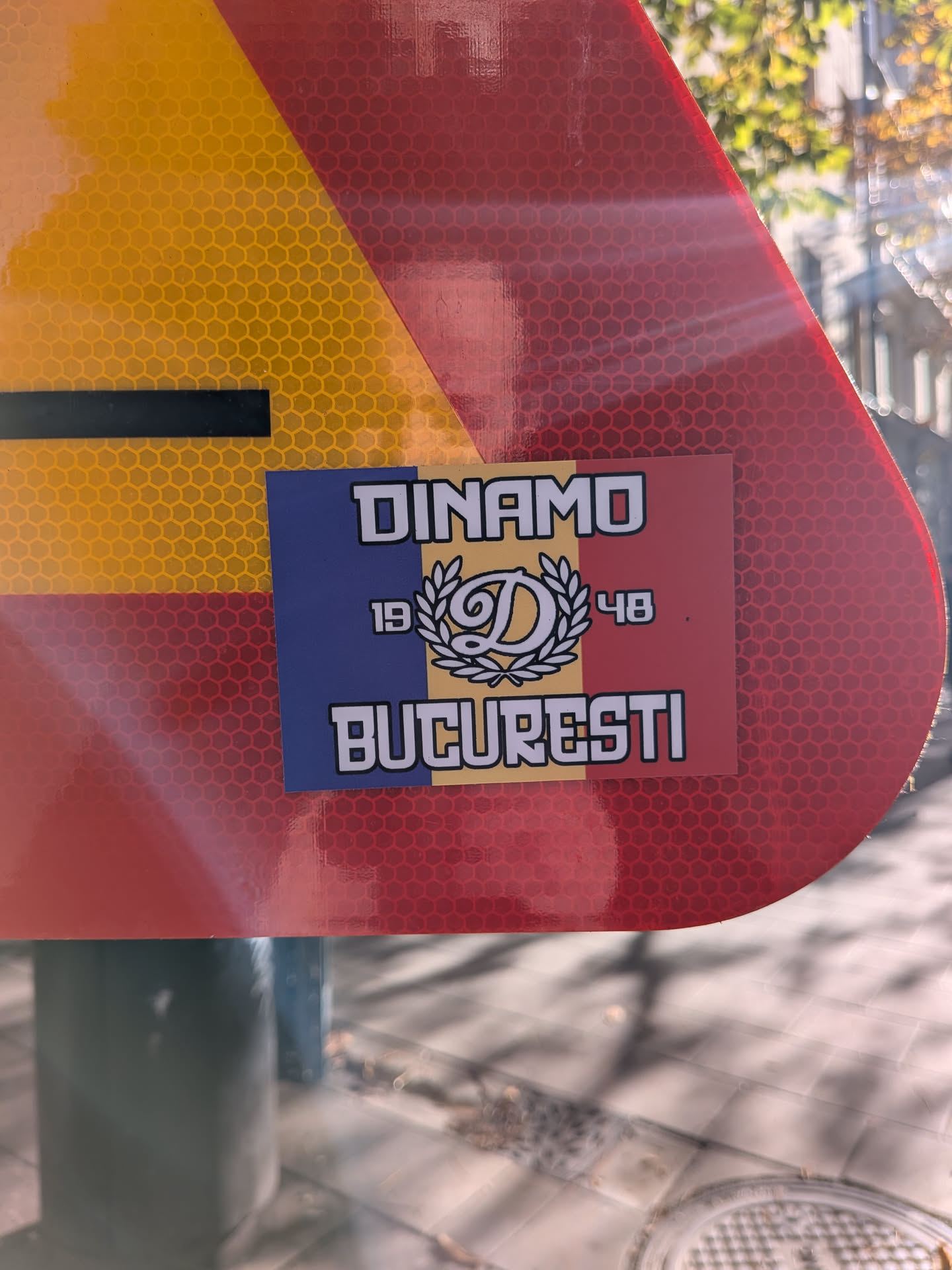 A rectangular vinyl sticker featuring the Romanian national tricolor stripes (blue, yellow, and red). Centered on top is the text 'DINAMO' in a bold, stylized white typeface with a black outline. The middle contains a laurel wreath framing a calligraphic letter 'D', flanked by the founding year '19' and '48'. Below it, the city name 'BUCURESTI' is printed in the same white block lettering. The sticker is adhered to a reflective red and yellow street sign.