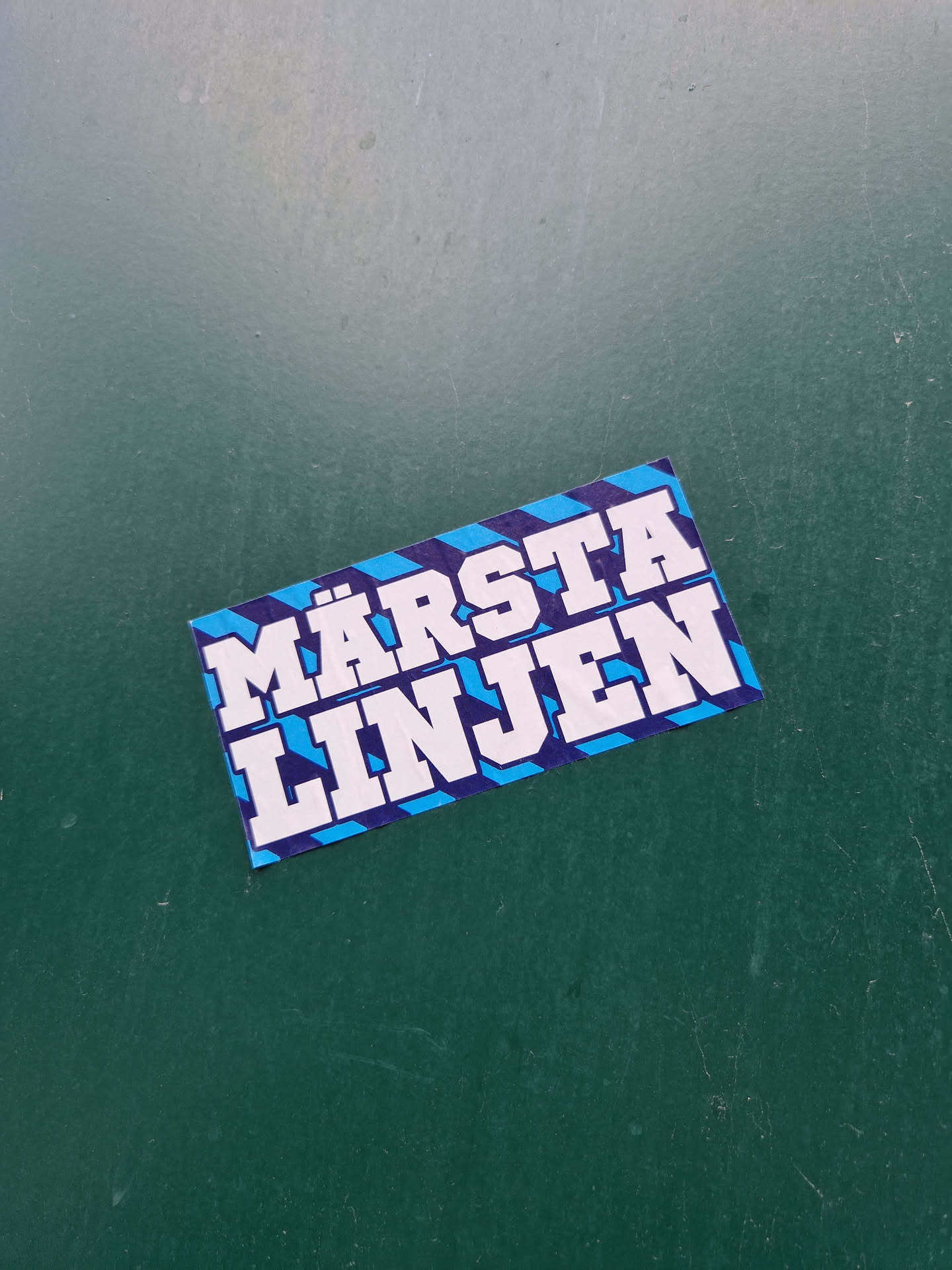 A blue and white sticker with the text "Märsta Linjen" on it. The text is in a bold, sans-serif font. The sticker has a diagonal stripe pattern.