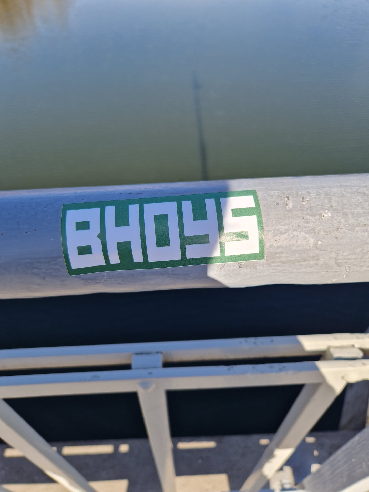 A green and white sticker with the word BHOYS in a stylized font is affixed to a metal railing near a body of water. The sticker appears to be part of a larger design or brand. 
