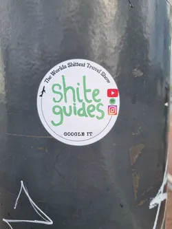 Round sticker with green text on a white background.  It says "Shîte guides" in a stylized font.  At the top it says "The World's Shittest Travel Show" in a smaller font. At the bottom it says "Google it". There are also social media icons for YouTube, Spotify and Instagram.
