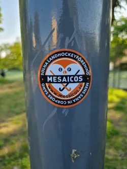 Round orange and white sticker with the text "Solna Landhockeyförening" around the perimeter. In the center is a heart with two crossed hockey sticks and the word "MESAICOS" below it. The website www.mesaicos.se is also printed on the sticker.