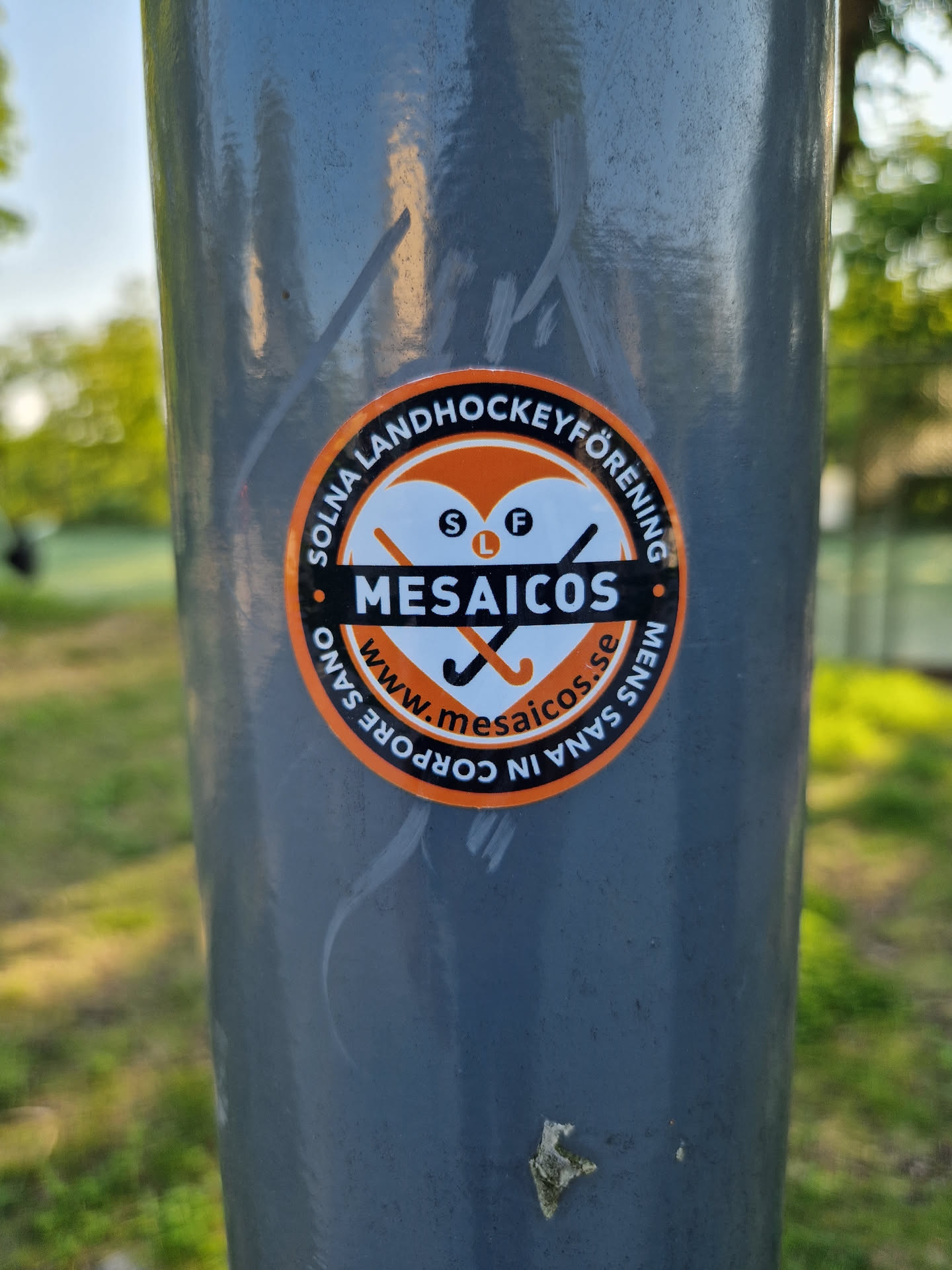 Round orange and white sticker with the text "Solna Landhockeyförening" around the perimeter. In the center is a heart with two crossed hockey sticks and the word "MESAICOS" below it. The website www.mesaicos.se is also printed on the sticker.