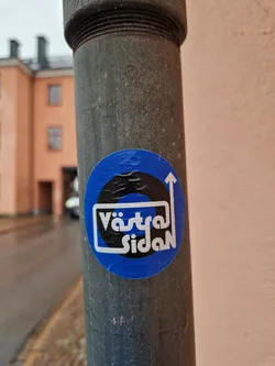 A blue circular sticker with black and white design. The text 'Västra Sidan' is visible within a white box.  An upward pointing arrow is also featured in the design.