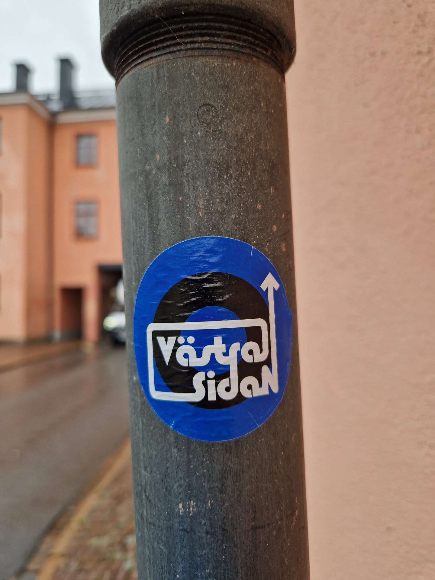 A blue circular sticker with black and white design. The text 'Västra Sidan' is visible within a white box.  An upward pointing arrow is also featured in the design.