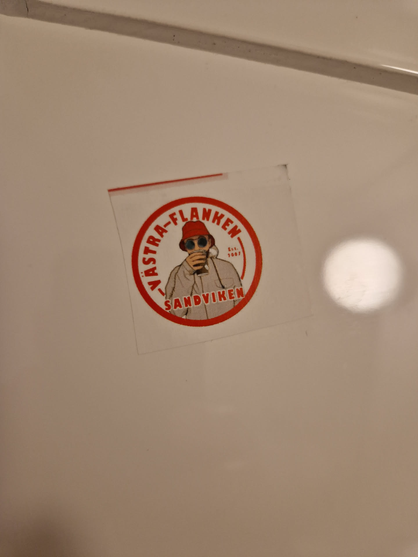 A round sticker with a red border and white background. It features an illustration of a person wearing a red bucket hat, sunglasses, and a light-colored jacket, holding a drink. The text "VäSTRA-FLANKEN" is written in a circle around the illustration, with "SANDVIKEN" written below.  The year "Est. 2007" is visible.