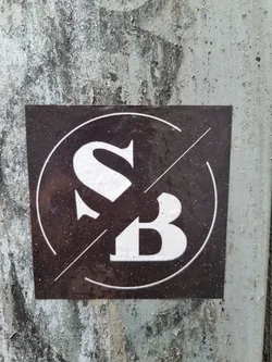 A dark-colored square sticker with a logo featuring the letters "SB" in a stylized font. The letters are white and are set within a circle with a diagonal line intersecting it. The sticker appears to be affixed to a weathered surface.