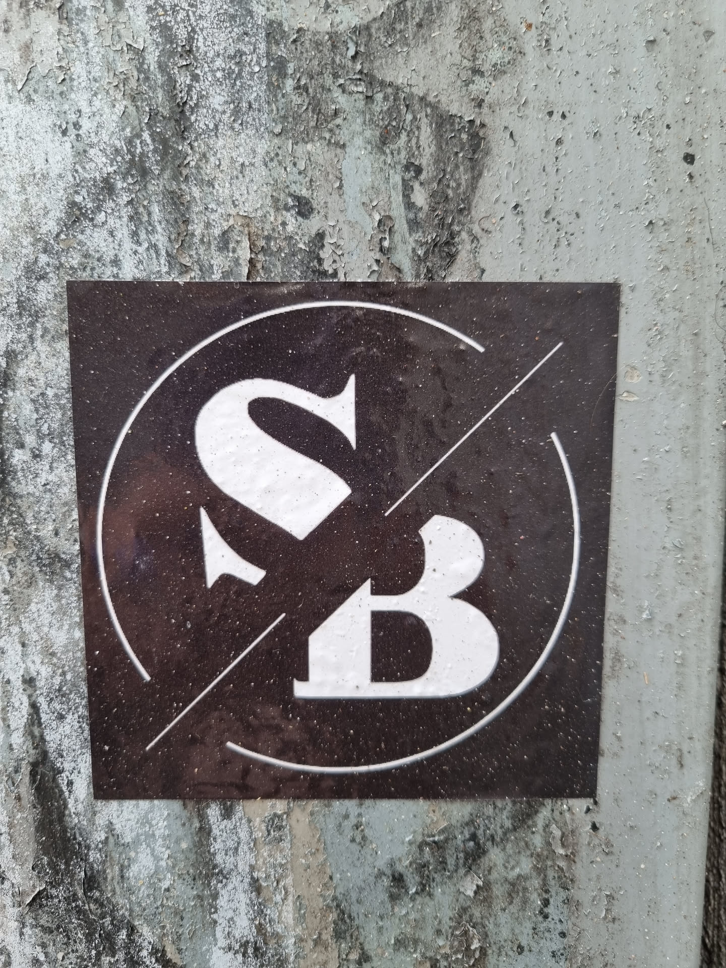A dark-colored square sticker with a logo featuring the letters "SB" in a stylized font. The letters are white and are set within a circle with a diagonal line intersecting it. The sticker appears to be affixed to a weathered surface.