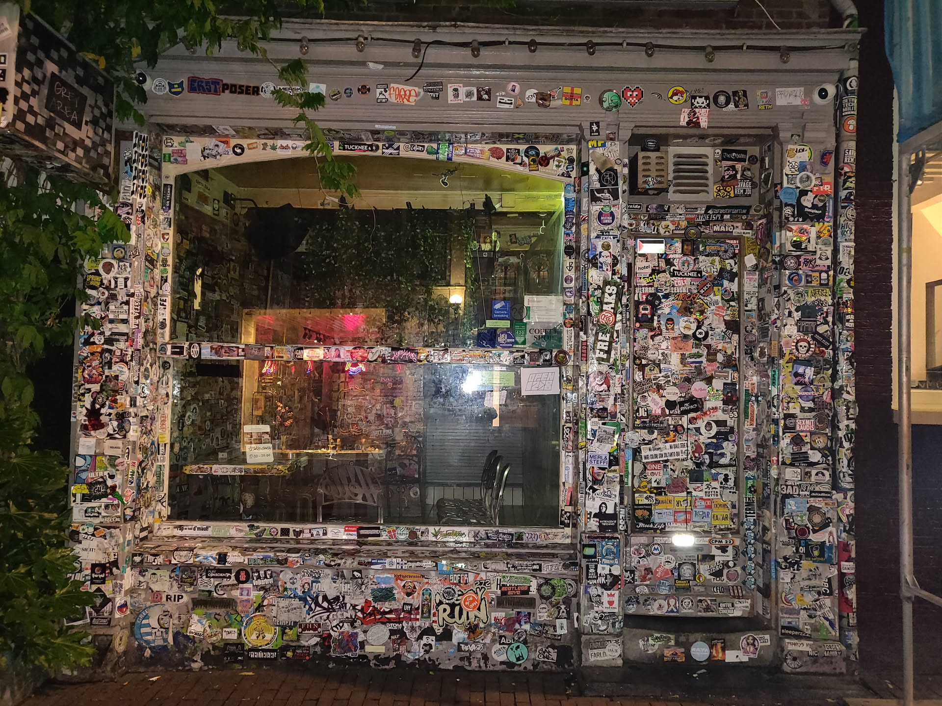 The image shows a building's exterior, completely covered in various stickers.  It appears to be a shop or bar, with a window showing a dimly lit interior. The stickers are diverse in design and style, creating a vibrant and layered visual texture.