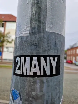 A black and white sticker with the text "2MANY" in a simple, bold font. The sticker is affixed to a metal pole. There are other, older stickers visible underneath.