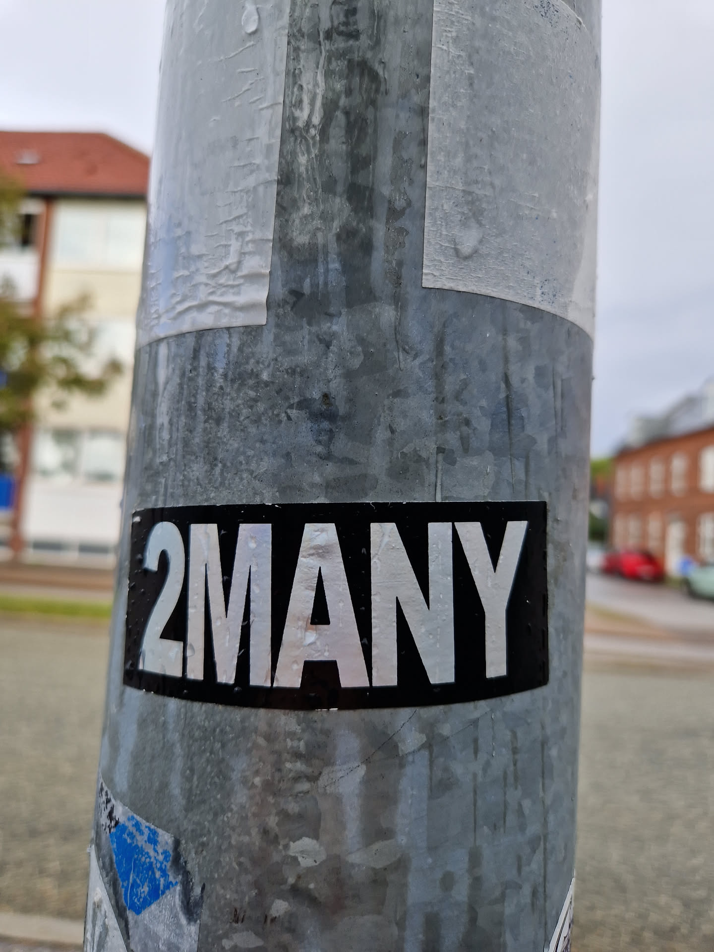 A black and white sticker with the text "2MANY" in a simple, bold font. The sticker is affixed to a metal pole. There are other, older stickers visible underneath.