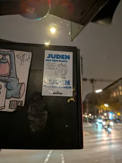 Street sticker A vertical rectangular sticker with a white background and blue and black printing, stuck onto a dark metal box outdoors. The top features bold blue text reading 'JUDEN AUF DEM PLATZ', followed by smaller explanatory text 'Jüdische Lebenswirklichkeiten und Antisemitismus im Fußball heute' next to a minimalist line illustration of a person wearing a coat and scarf. Across the lower half, faded blue lettering reads 'JUDEN AUF DEN RÄNGEN'. At the bottom center, a small black logo contains the name 'DIE WERKSTATT'. Water droplets are visible on its surface.