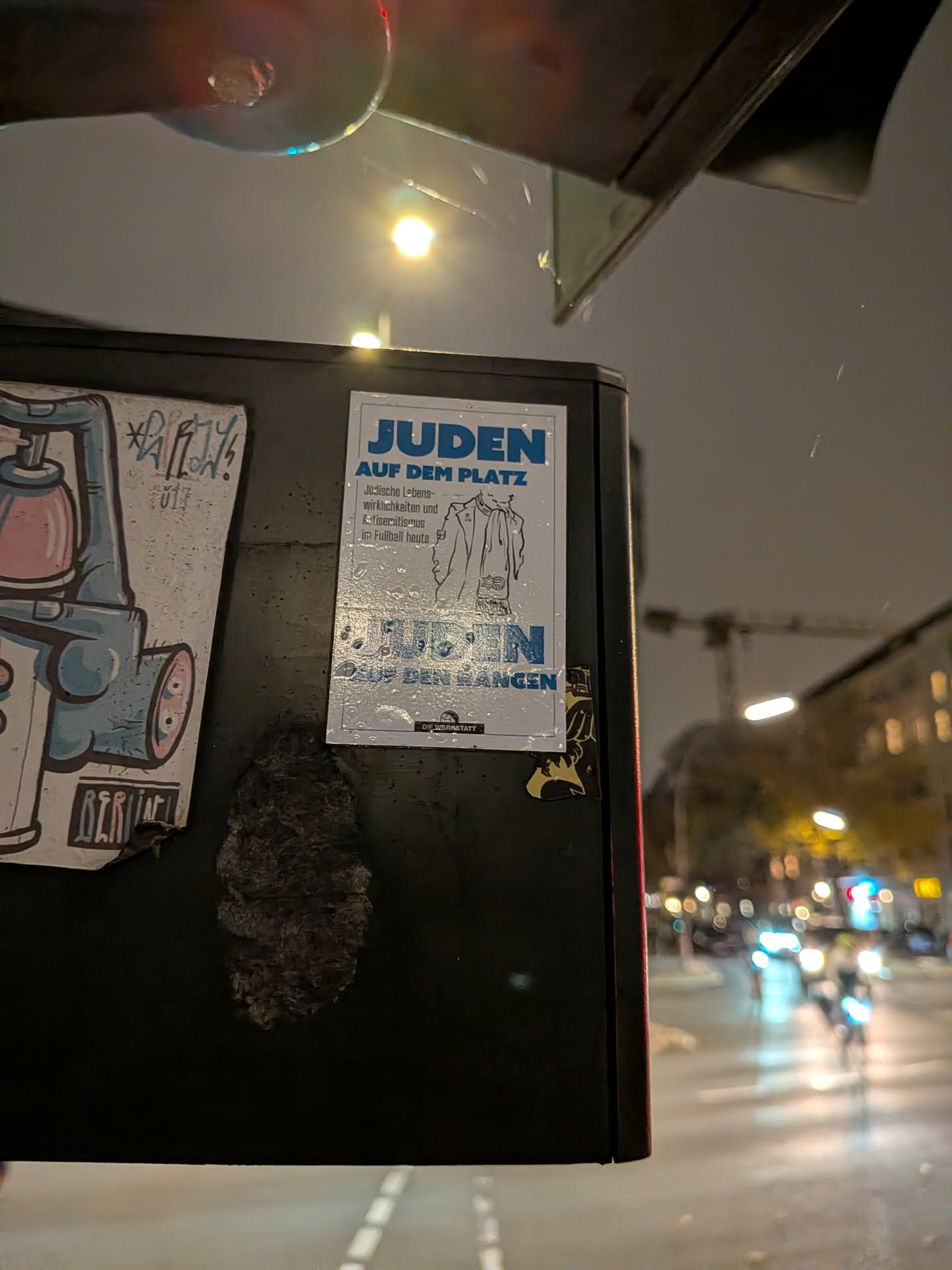 A vertical rectangular sticker with a white background and blue and black printing, stuck onto a dark metal box outdoors. The top features bold blue text reading 'JUDEN AUF DEM PLATZ', followed by smaller explanatory text 'Jüdische Lebenswirklichkeiten und Antisemitismus im Fußball heute' next to a minimalist line illustration of a person wearing a coat and scarf. Across the lower half, faded blue lettering reads 'JUDEN AUF DEN RÄNGEN'. At the bottom center, a small black logo contains the name 'DIE WERKSTATT'. Water droplets are visible on its surface.