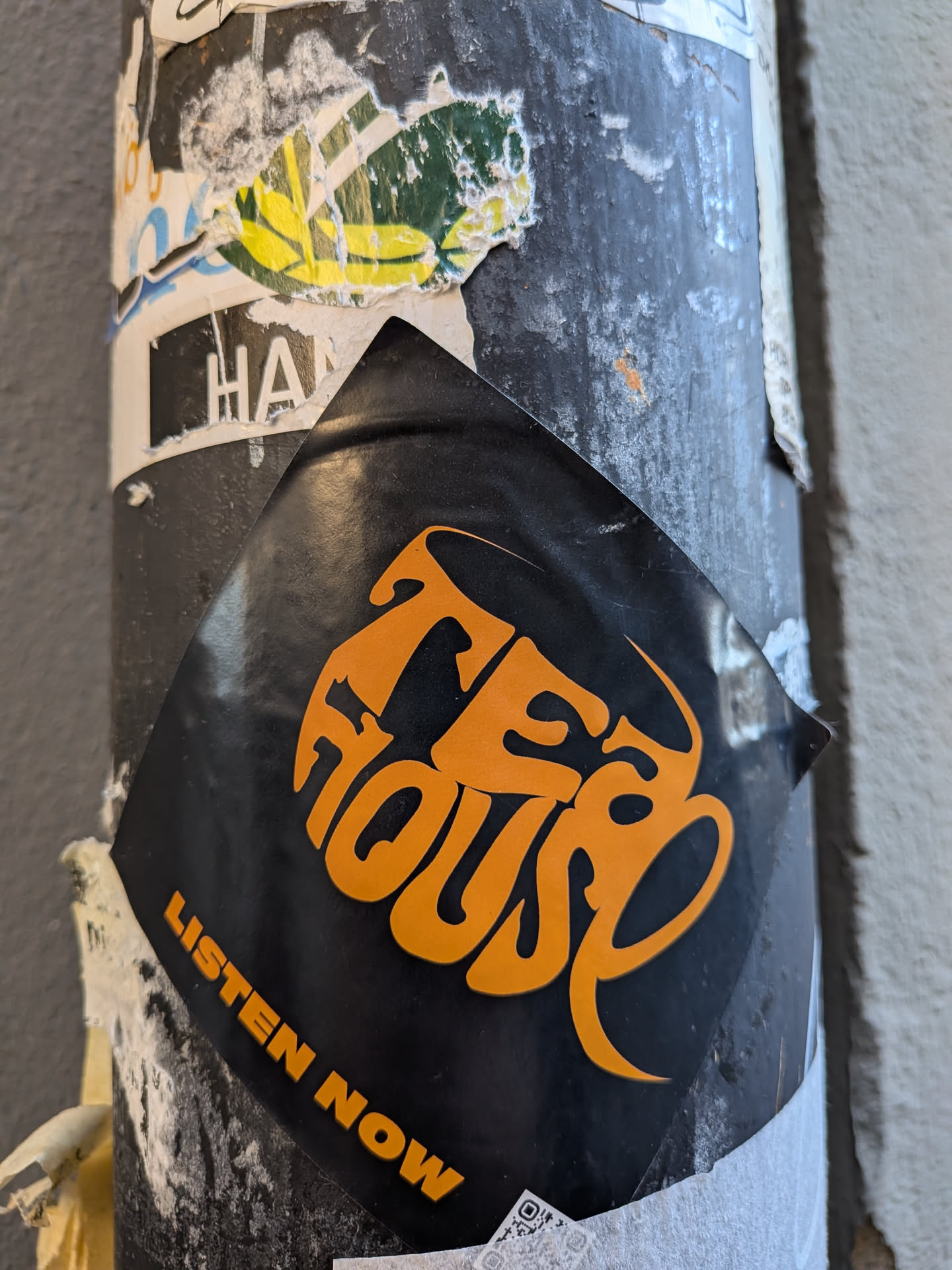 A diamond-shaped sticker with a matte black background. The central design features the words 'TEA HOUSE' in a highly stylized, flowing, interconnected orange font that evokes a retro or psychedelic aesthetic. Below this main text, along the bottom left edge, the phrase 'LISTEN NOW' is written in a simpler, all-caps orange sans-serif font. The sticker shows signs of wear, with slight curling at the edges and minor surface imperfections, indicating it's a physical sticker adhered to a textured, dark pole.
