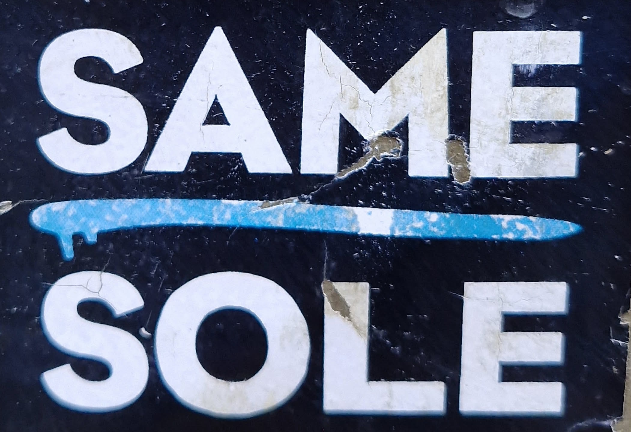 A worn sticker with the text SAME over SOLE, separated by a light blue line. The sticker has a dark background and shows signs of wear and tear.