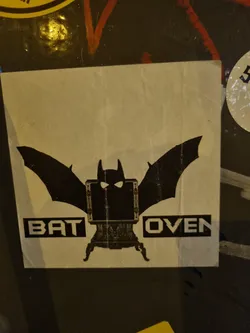 A sticker featuring a bat-shaped silhouette incorporating an old-fashioned oven, with the words "BAT OVEN" written underneath. The style is dark and slightly quirky.