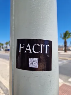 Black sticker with the word FACIT in white capital letters and a QR code in the center. The sticker is attached to a light-grey pole outdoors.
