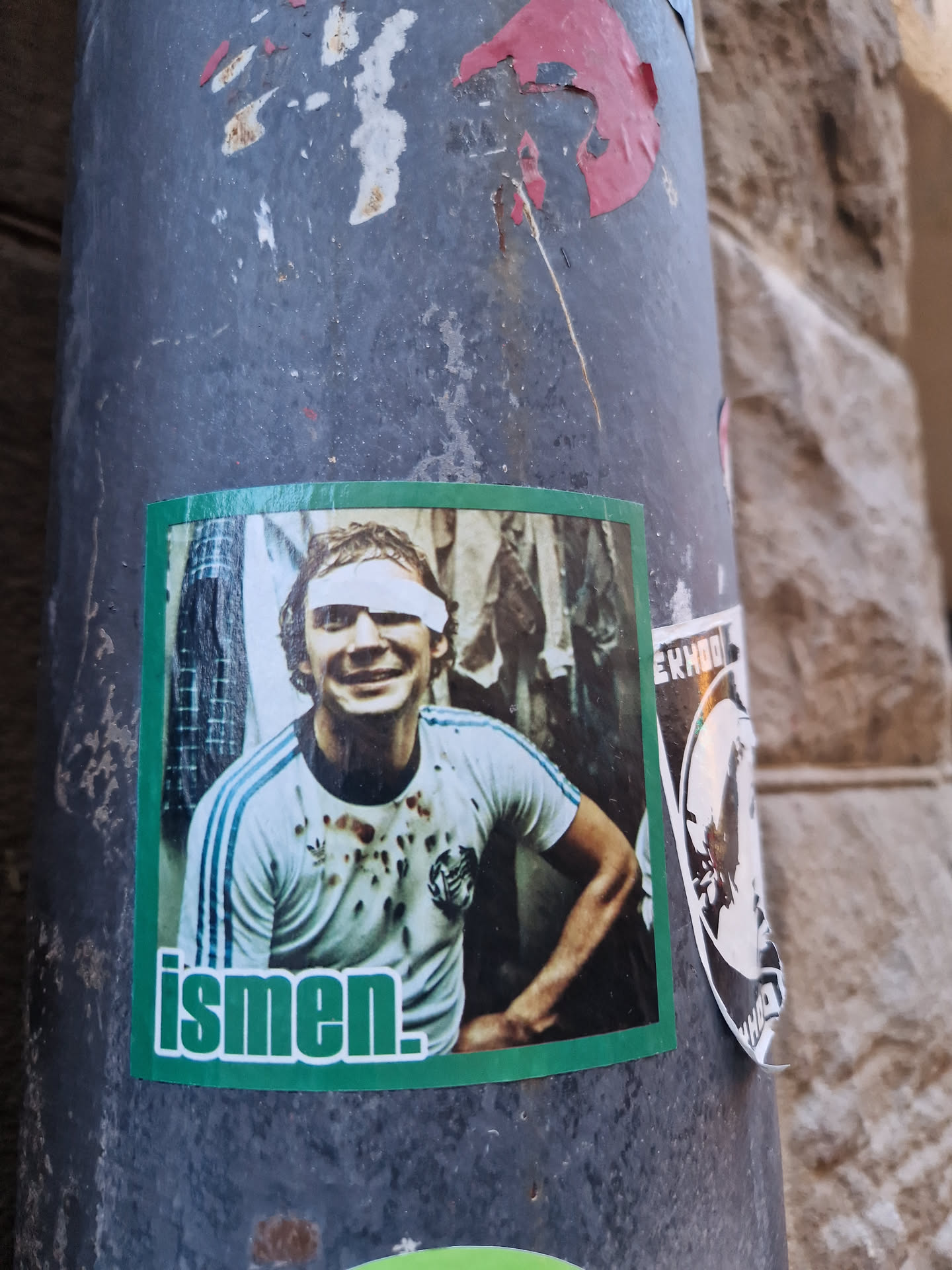 A green sticker with a picture of a man with a white headband. The man is wearing a white short sleeved shirt with three stripes on the shoulders. The background of the photo is blurry but appears to be in a changing room. The word 'ismen.' is written in green letters at the bottom of the sticker. The sticker is affixed to a gray metal pole with other stickers and markings.