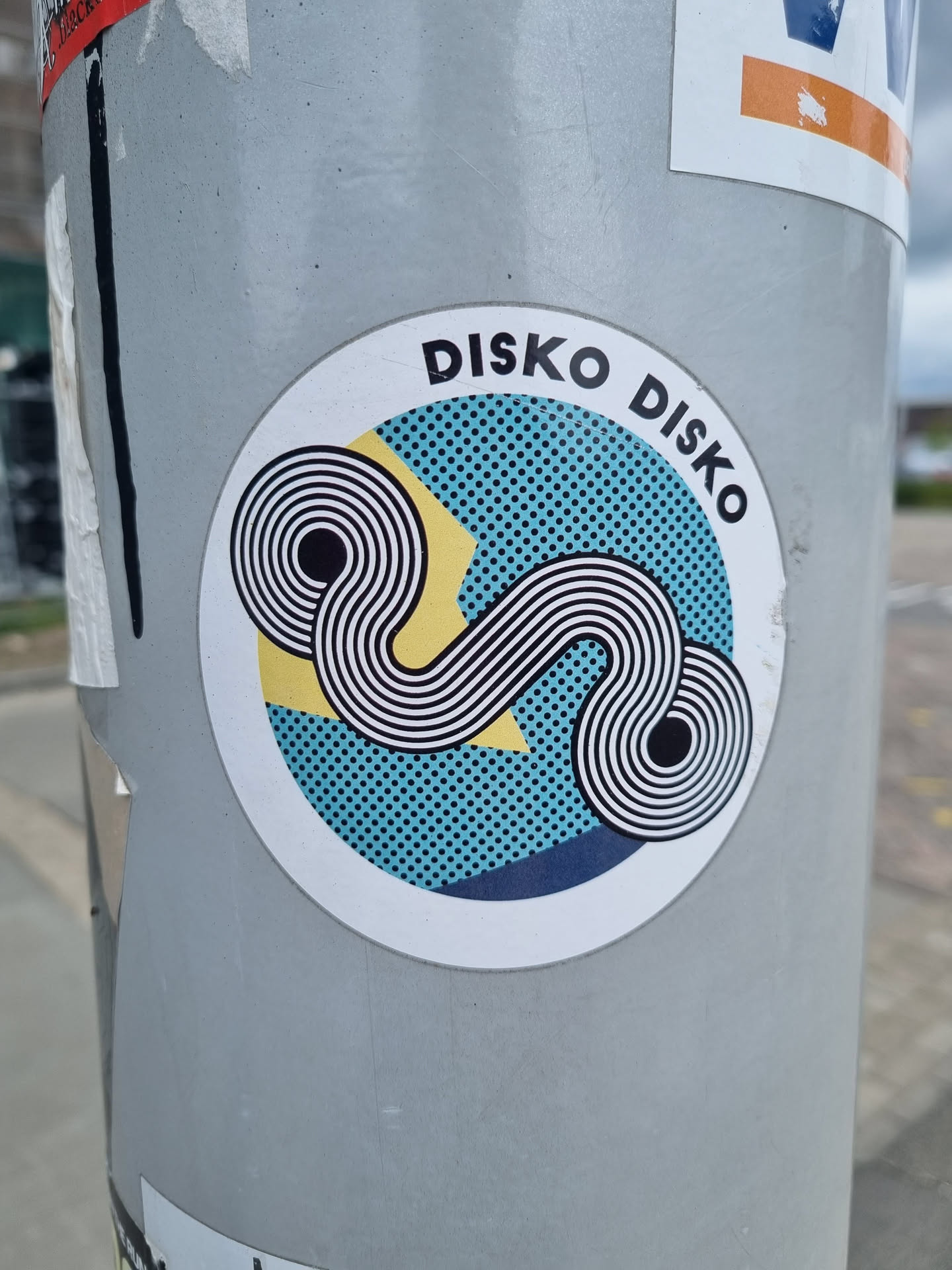 Round sticker with the text DISKO DISKO. It features an abstract design with teal, yellow, and white colors, along with black concentric circles. The style is reminiscent of 70s or 80s graphic design.