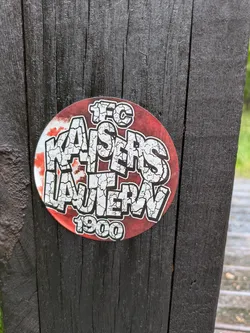 A circular sticker featuring the name of a football club, '1. FC Kaiserslautern', in large, white, cracked-texture letters with a black outline against a red background. The text '1. F.C.' is at the top in a similar style, and '1900' is at the bottom. The sticker is applied to a dark, vertically grained wooden surface, possibly a fence post or railing, which shows signs of weathering.