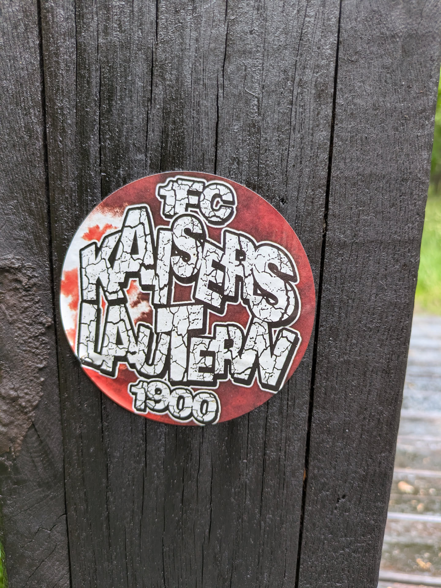 A circular sticker featuring the name of a football club, '1. FC Kaiserslautern', in large, white, cracked-texture letters with a black outline against a red background. The text '1. F.C.' is at the top in a similar style, and '1900' is at the bottom. The sticker is applied to a dark, vertically grained wooden surface, possibly a fence post or railing, which shows signs of weathering.