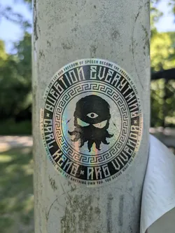 A holographic sticker featuring an eye with a tentacle-like body, surrounded by text encouraging critical thinking and independence. The sticker is affixed to a metal pole.