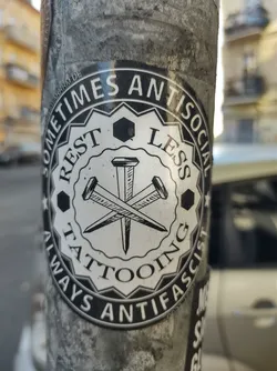 A black and white sticker with a circular design. The center features two crossed nails over a hexagon. The text around the circle says: "Sometimes Antisocial Restless Tattooing Always Antifascist".
