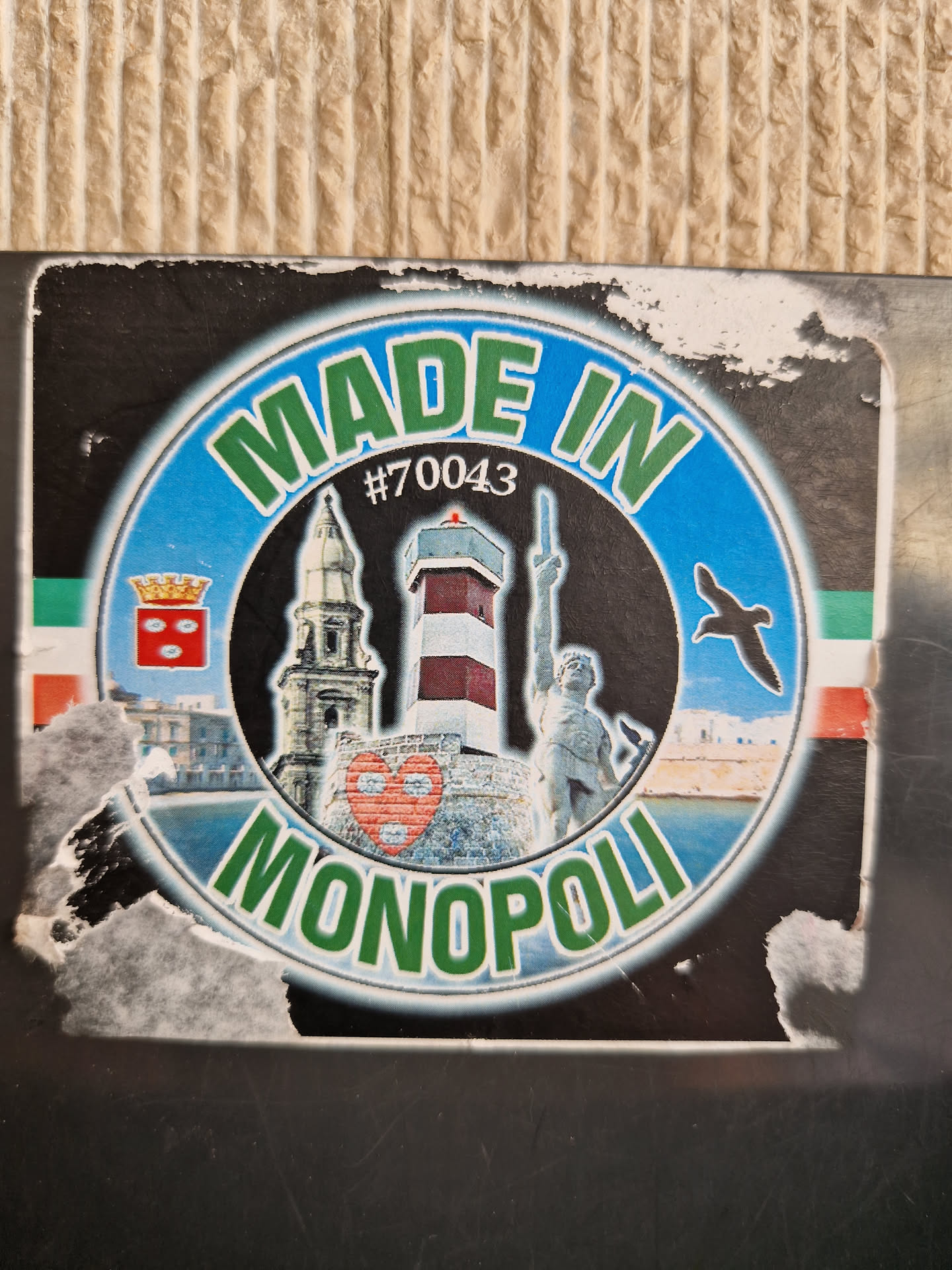 A sticker featuring landmarks of Monopoli, Italy. The sticker has a circular design with the text "Made in Monopoli" in green lettering. Inside the circle are images of a lighthouse, a church, and a statue, along with a small red heart. There is a small code #70043 visible on the sticker.