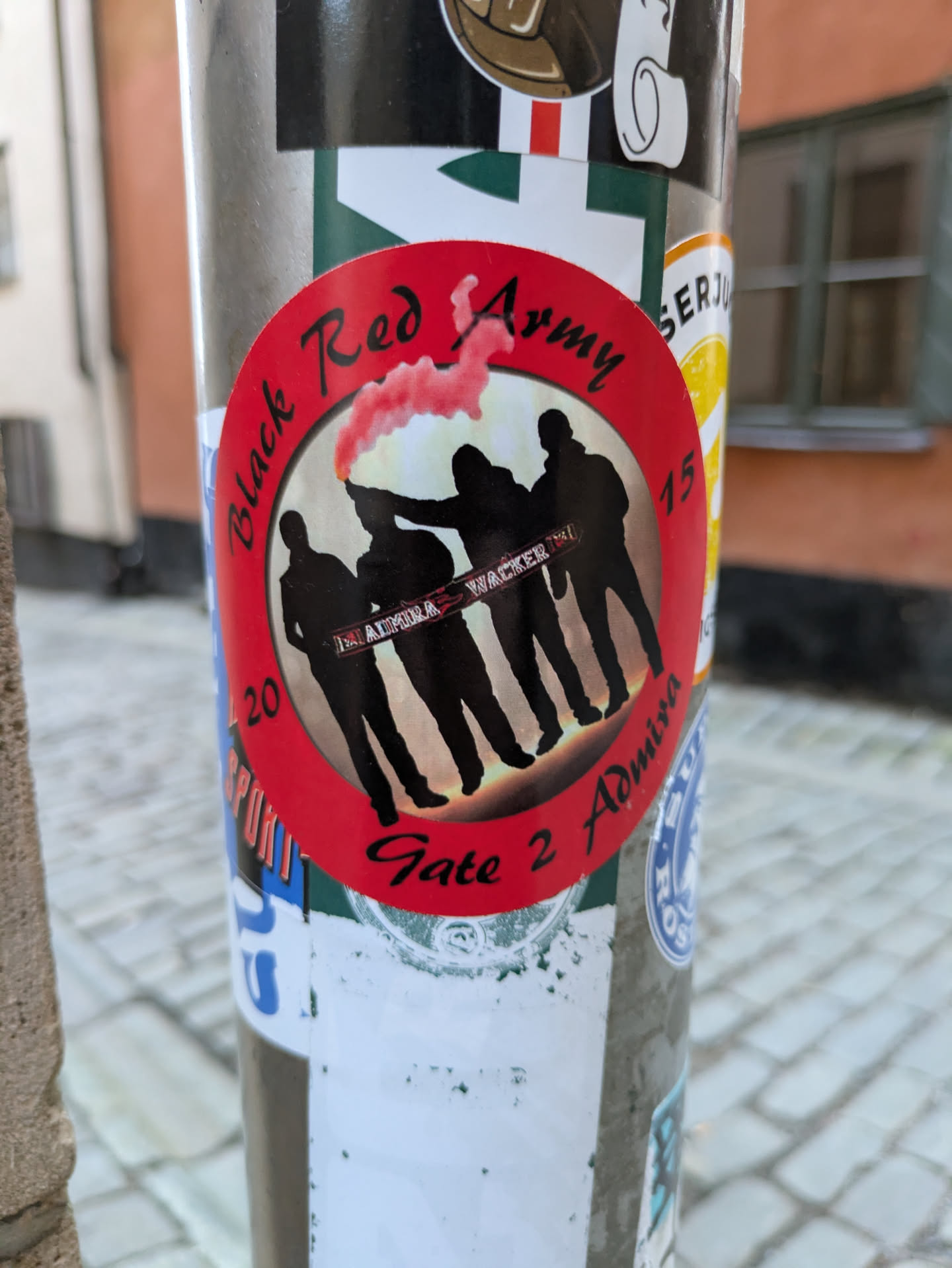 Round sticker with a red background featuring silhouettes of people against a backdrop of a red flare or smoke. Text on the sticker includes 'Black Red Army', 'Gate 2 Admission', and partially visible text that seems to say 'Arena Wacker'. The numbers '20' and '15' are visible on either side of the design. 