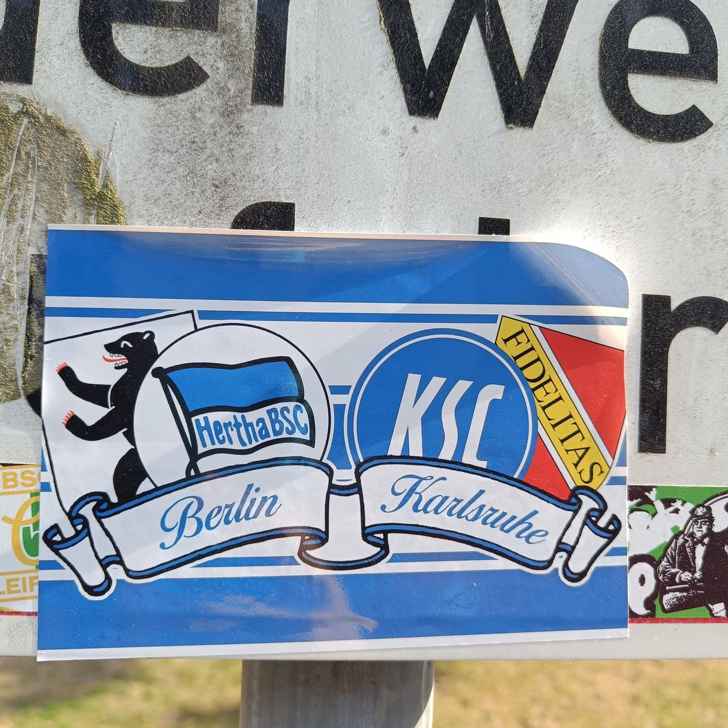A sticker showing the logos of Hertha BSC Berlin and Karlsruher SC, with their city names written on banners underneath. The design is in blue and white, featuring the Berlin bear and KSC's Fidelitas.