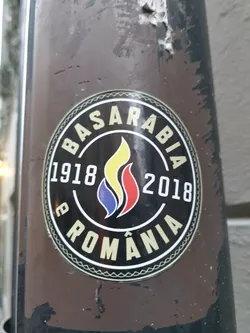 A round sticker commemorating the union of Basarabia with Romania in 1918. The sticker features the years 1918 and 2018, a stylized flame in the colors of the Romanian flag, and the text "Basarabia" and "România" around the flame. The sticker is affixed to a brown pole.
