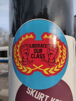 A round sticker with a red and yellow design featuring two fists in chains, gears, and laurel wreaths. The text 'Liberate Our Class' is prominently displayed.