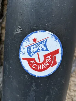 A worn FC Hansa Rostock sticker on a dark surface. The sticker features the team's logo, which includes a griffin on a ship. The colors are primarily blue, red and white.