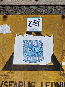 A sticker featuring the text 'Terr Ballin' and an illustration of people sitting on top of a pile of skulls. The sticker is on a yellow sign. There's also a smaller sticker above it with the text 'Eonity'.