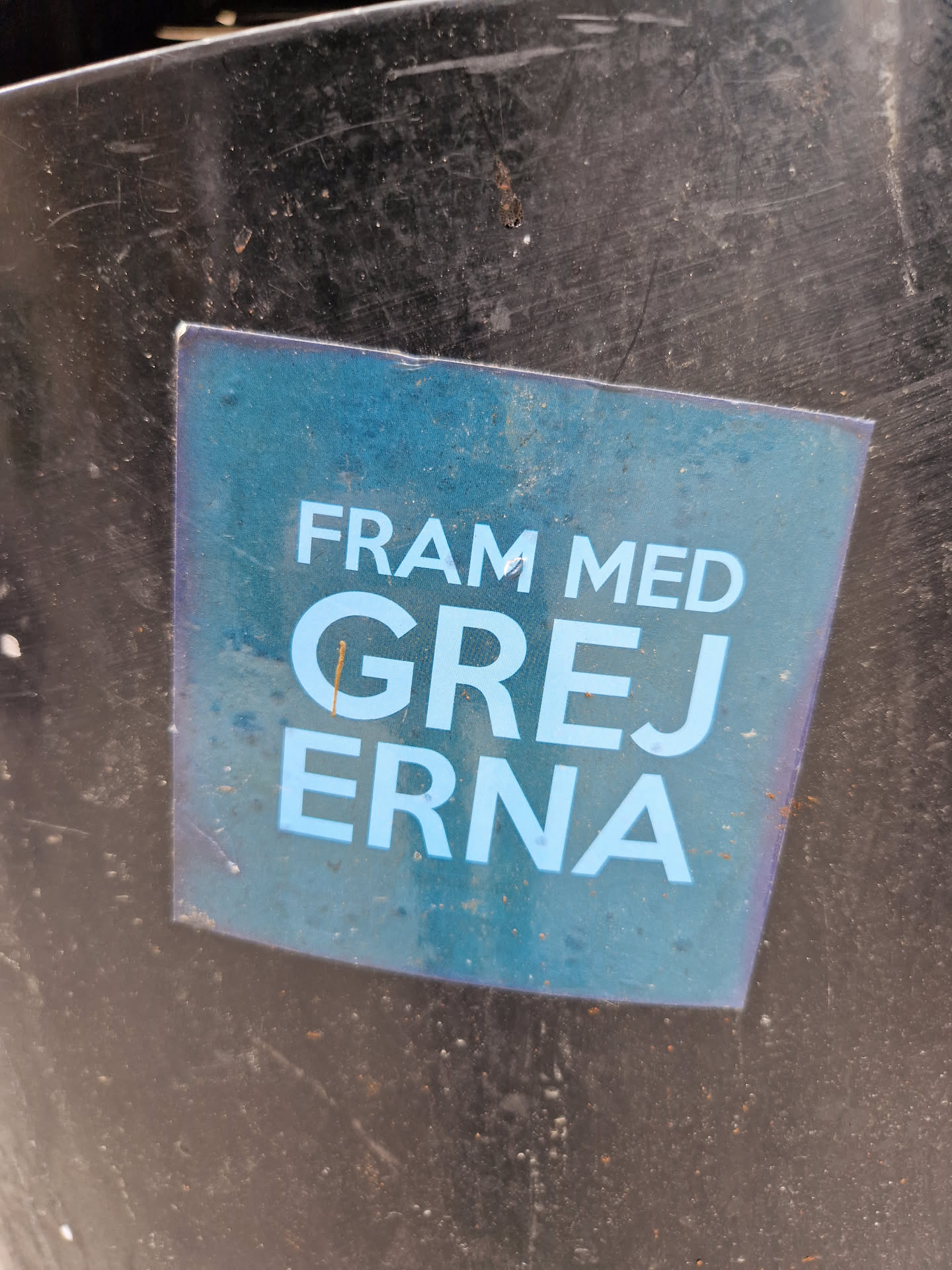 A blue sticker on a dark surface with the text "Fram med grejerna" which translates to "Bring out the things" or "Get the stuff out".