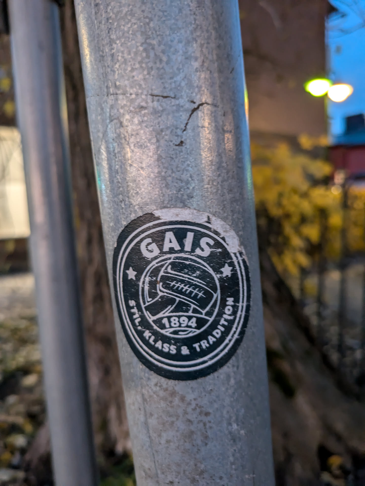 A black and white sticker featuring the logo of the Swedish football club GAIS (Göteborgs Atlet- och Idrottsförening). The logo includes the club's name, founding year (1894), and the motto "Stil, Klass & Tradition" (Style, Class & Tradition).  The sticker is affixed to a metal pole.