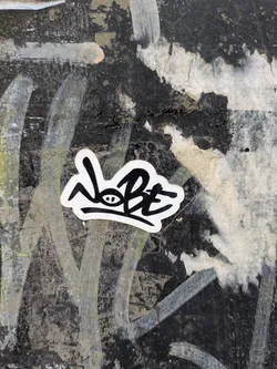A black and white sticker featuring the stylized text "NOBE" is affixed to a textured surface that appears weathered and worn. The background consists of a mix of dark and light patches, possibly from paint or residue.
