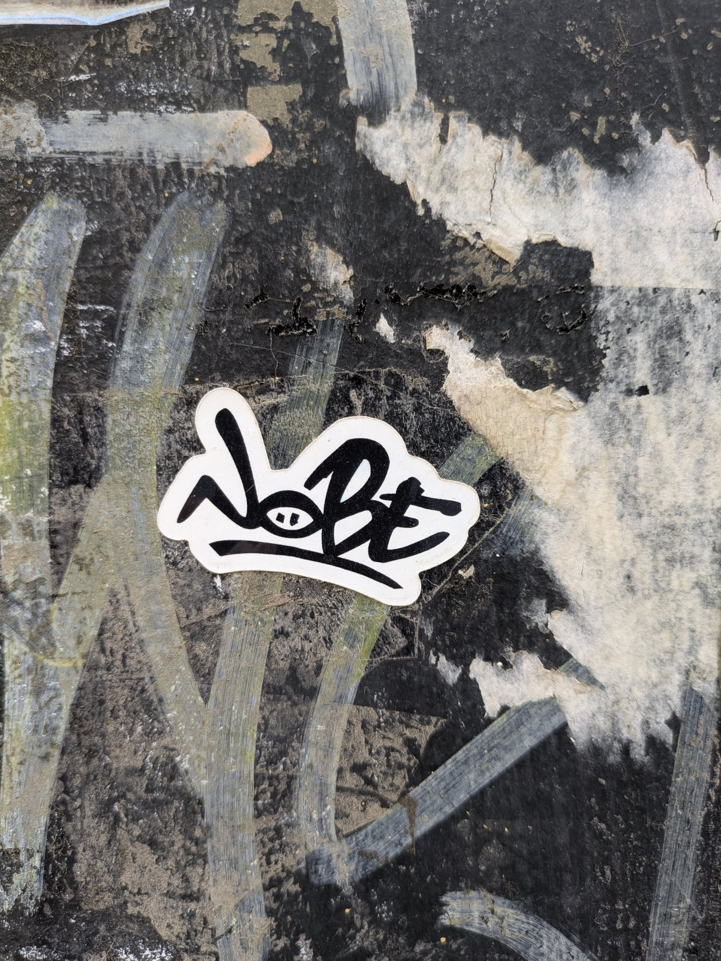 A black and white sticker featuring the stylized text "NOBE" is affixed to a textured surface that appears weathered and worn. The background consists of a mix of dark and light patches, possibly from paint or residue.