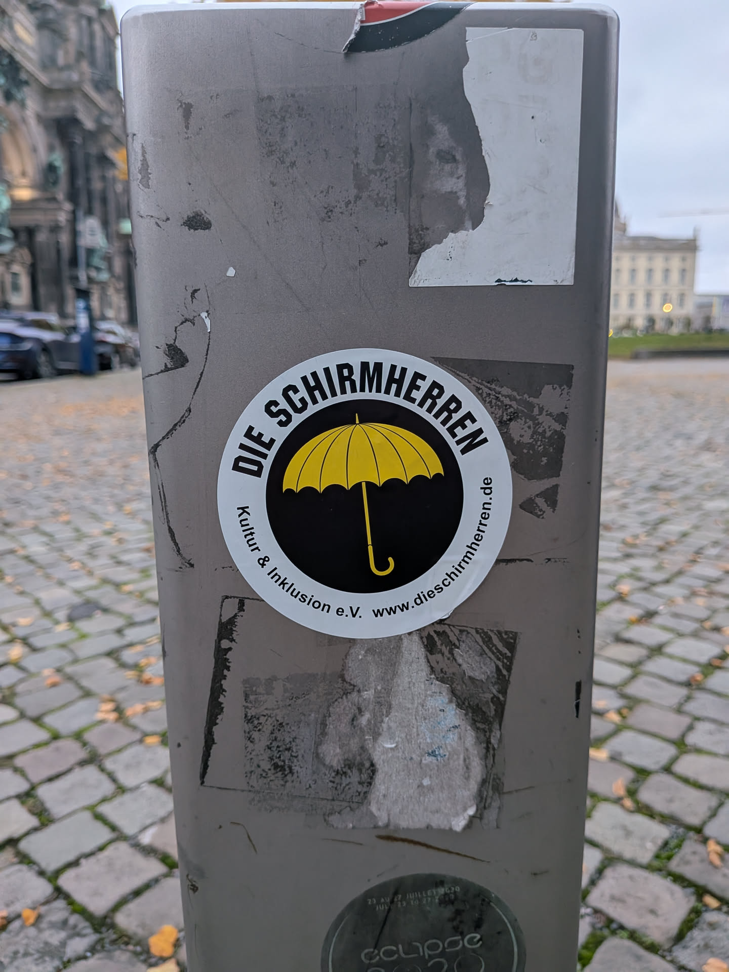 A round sticker with a white outer ring and a black circular center. Inside the black circle, there is a graphic illustration of an open yellow umbrella with a curved handle. Arching over the top in bold black capital letters is the text 'DIE SCHIRMHERREN'. Curving along the bottom within the white margin is the smaller text 'Kultur & Inklusion e.V. www.dieschirmherren.de'.