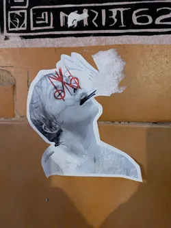 This sticker presents a black and white image of a person looking up and to the left. The portrait has red artistic marks around the eyes. The person is seemingly smoking a cigarette. The sticker is placed on a wall.