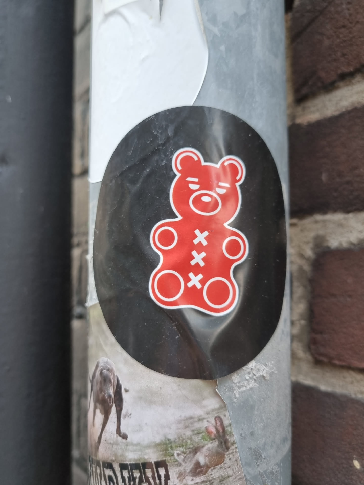 A black and red gummy bear sticker with a slightly annoyed expression. The sticker is oval-shaped and is placed on a grey metal pole.