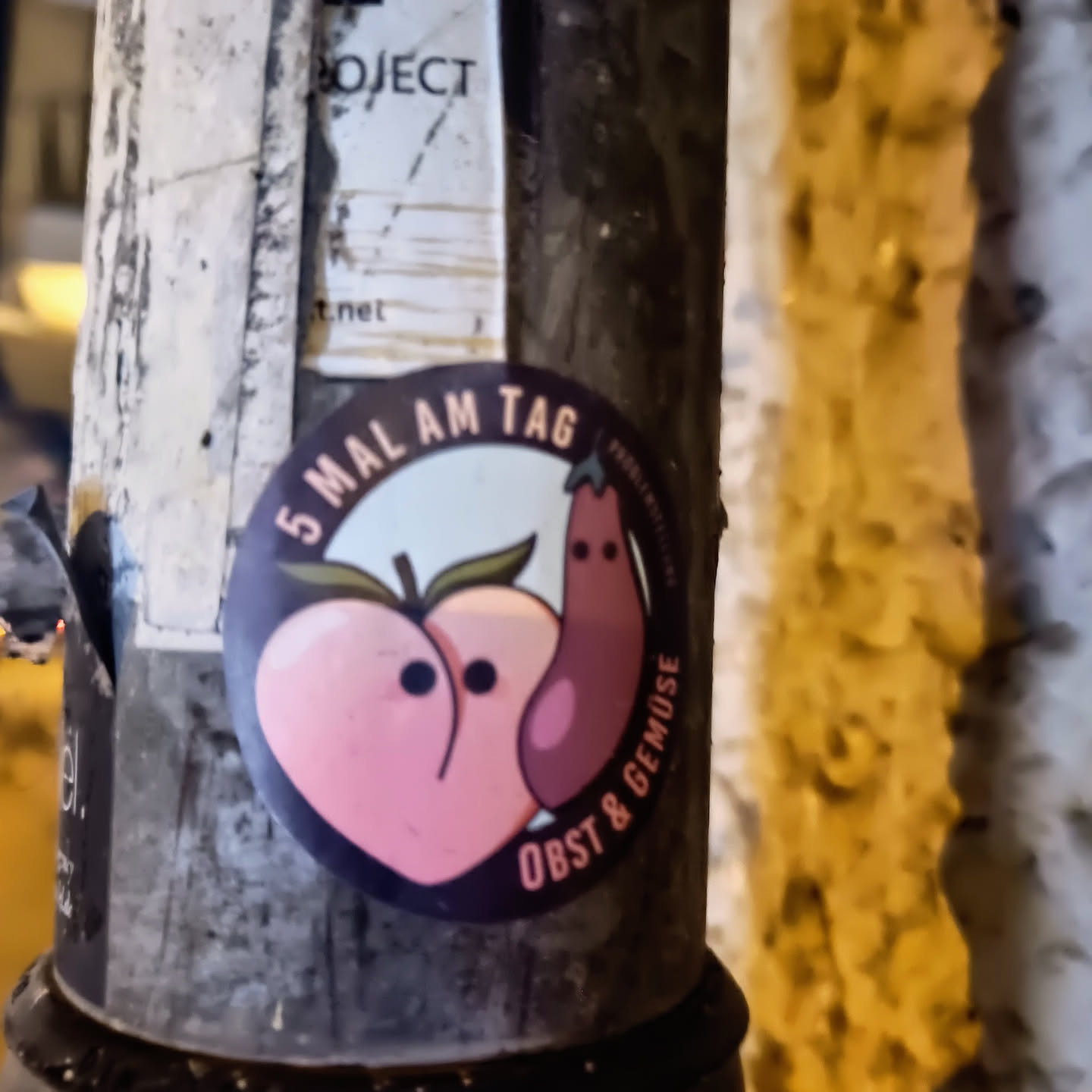 A round sticker featuring a peach and an eggplant with cute faces. The text on the sticker says "5 Mal am Tag Obst & Gemüse" which translates to "5 times a day fruit & vegetables".