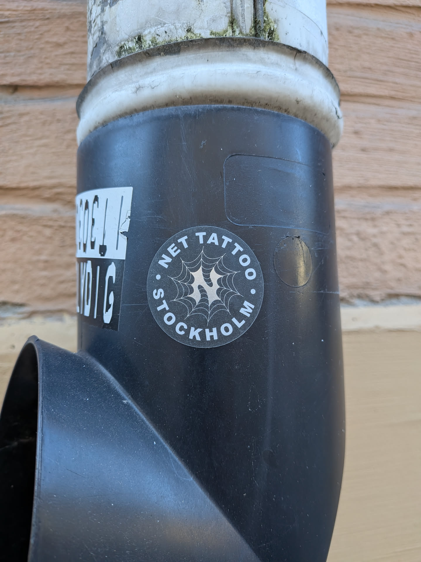 A circular monochrome sticker applied to a black plastic drainpipe. The sticker has a black background framed by curved white text reading 'NET TATTOO' across the top and 'STOCKHOLM' across the bottom, separated by white dots on the sides. At the center is an intricate white spiderweb illustration with a sharp, stylized letter 'N' integrated into its core.