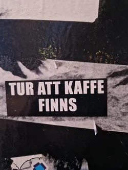 A black and white sticker with the text "TUR ATT KAFFE FINNS" (Swedish for "Time for coffee exists") on it. The sticker is attached to a dark surface. 