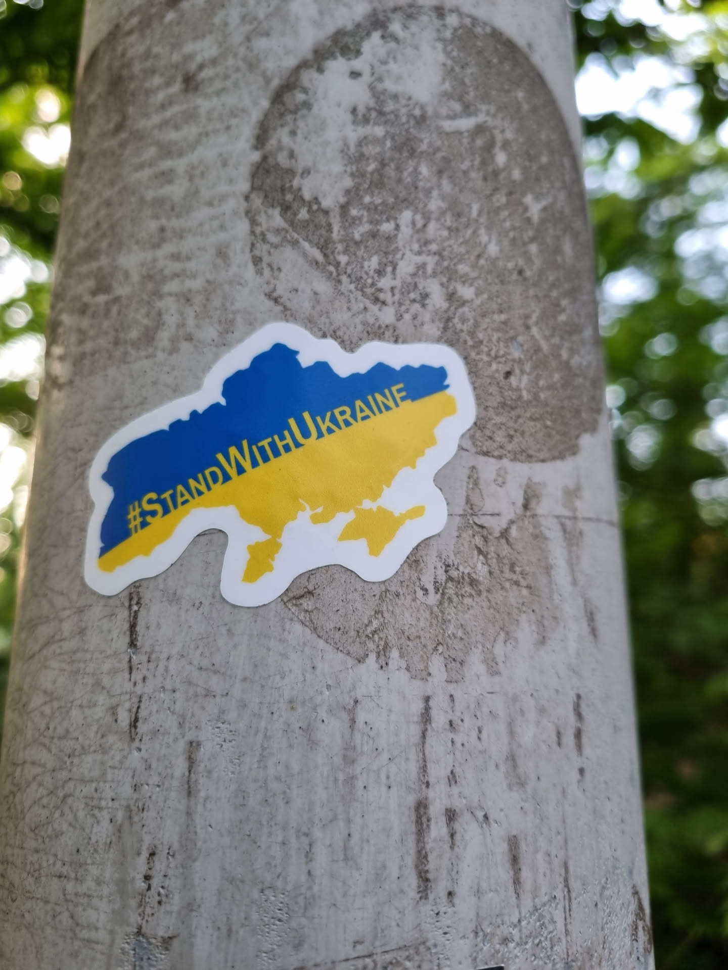 A sticker expressing solidarity with Ukraine, featuring a map of the country in its national colors (blue and yellow).  The sticker is affixed to a gray pole outdoors.