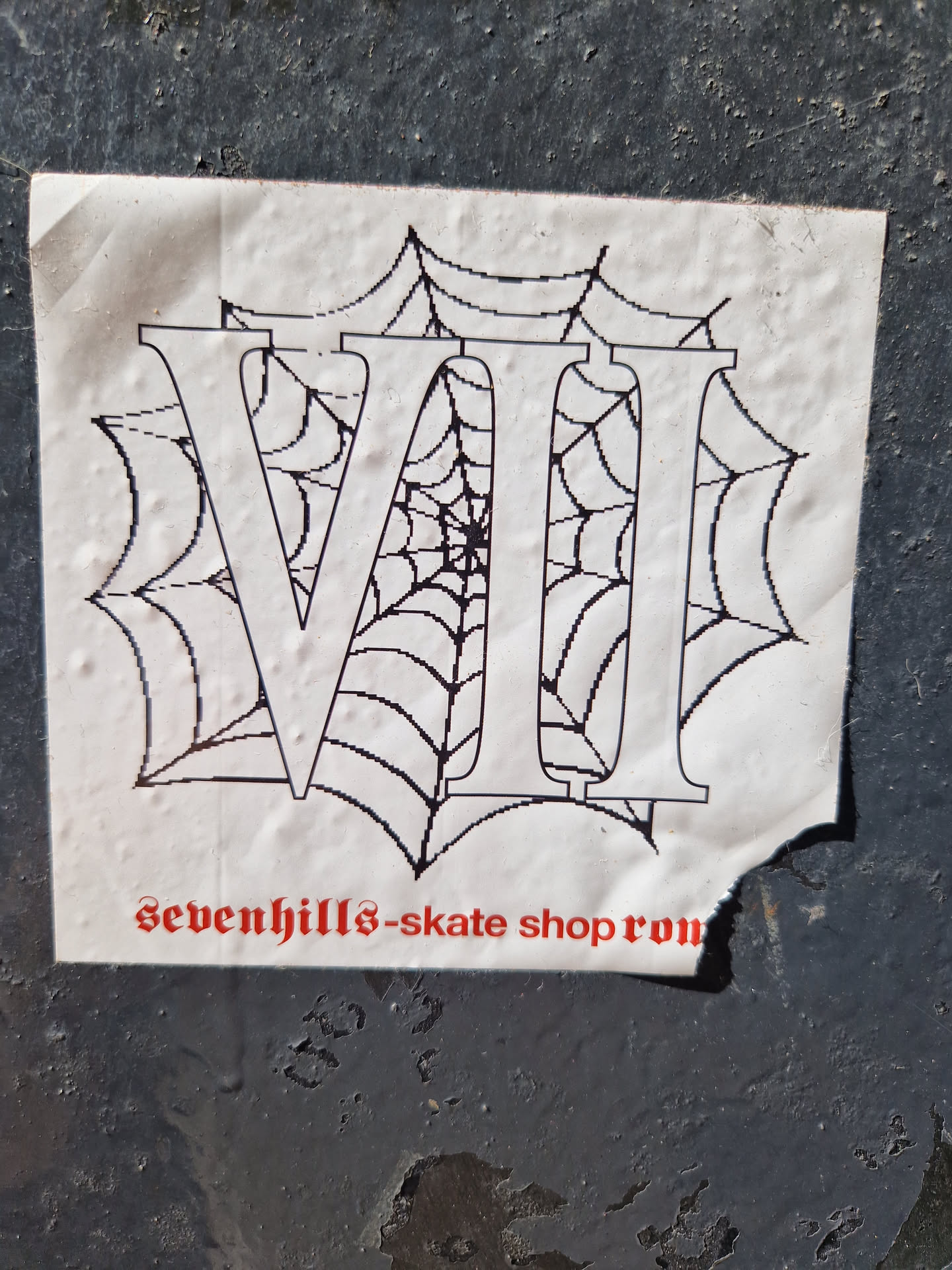 A sticker featuring the Roman numerals VII (7) overlaid on a spiderweb design. The text 'sevenhills-skate shop ron' is visible at the bottom. The sticker appears to be placed on a dark gray surface with some texture.