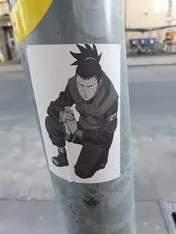 This sticker depicts Shikamaru Nara, a character from the popular anime and manga series Naruto, in a crouched, contemplative pose. The art style is grayscale, giving it a somewhat vintage feel. The sticker is affixed to a metal pole outdoors.