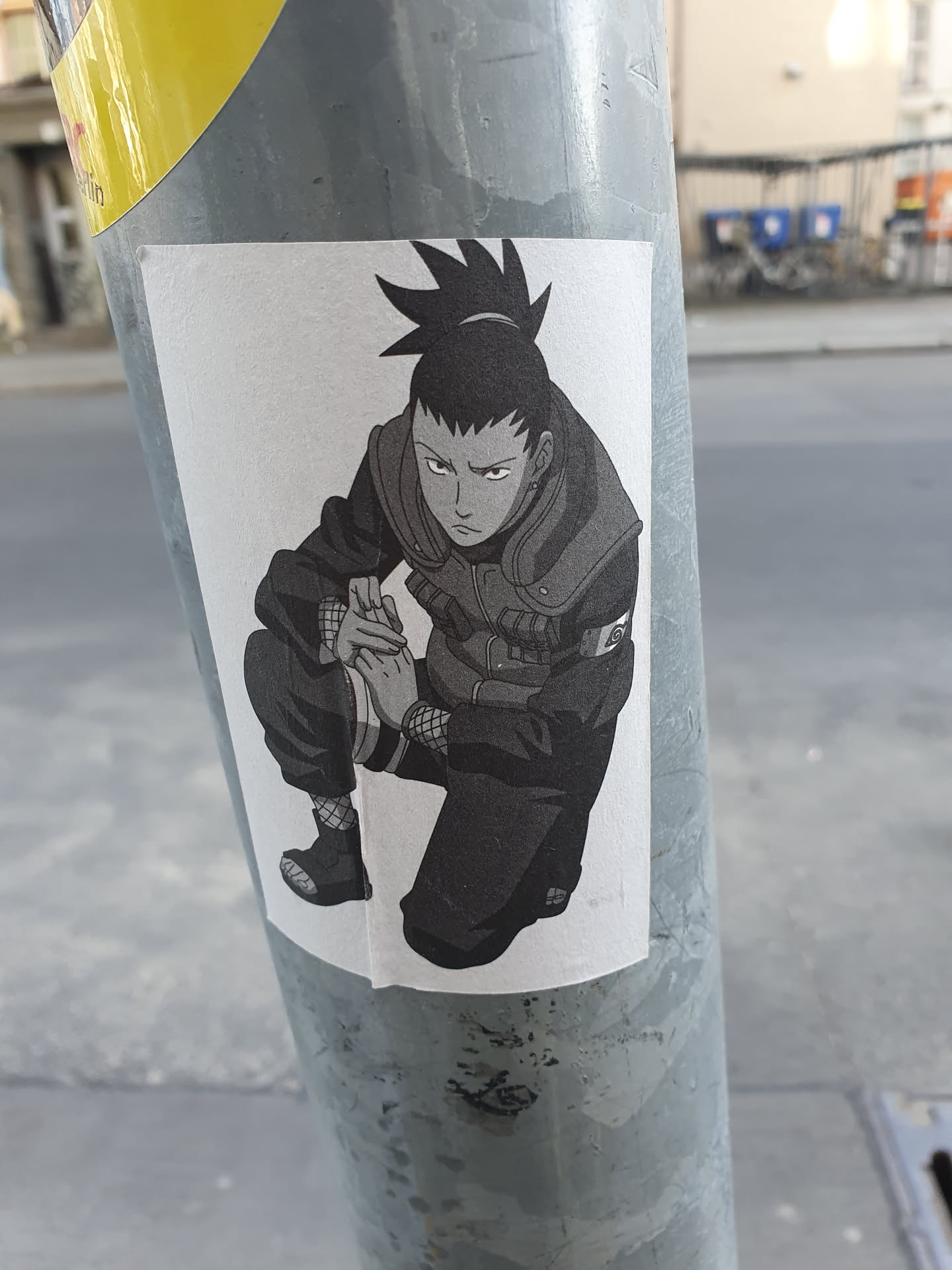 This sticker depicts Shikamaru Nara, a character from the popular anime and manga series Naruto, in a crouched, contemplative pose. The art style is grayscale, giving it a somewhat vintage feel. The sticker is affixed to a metal pole outdoors.