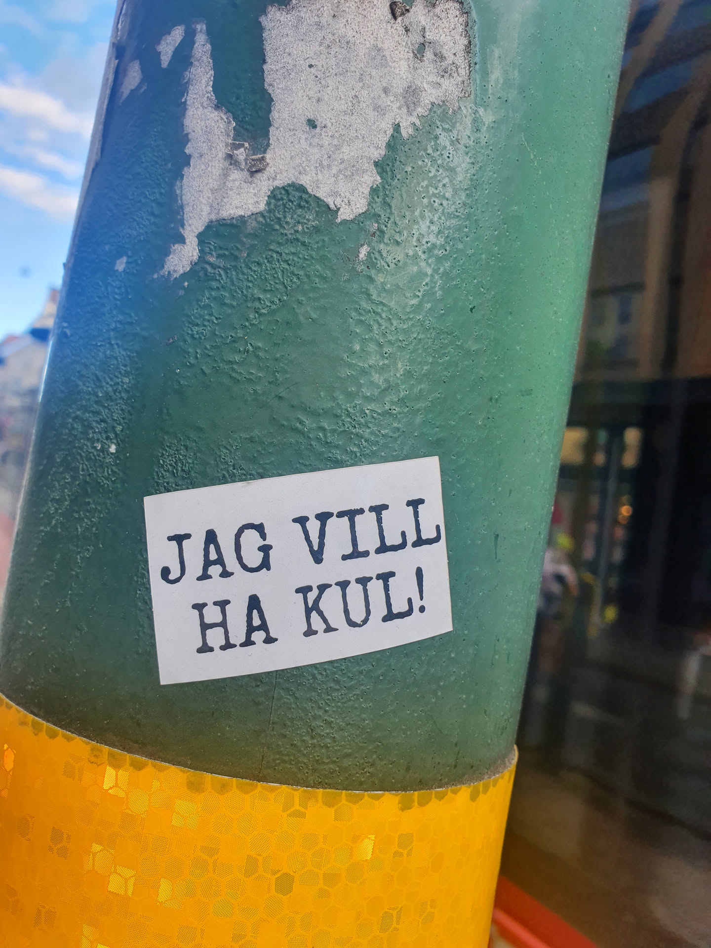 A simple sticker with the text "Jag vill ha kul!" (I want to have fun!) on it, placed on a green pole.