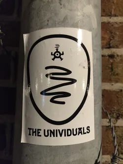 A white oval sticker with black outlines features a small symbol that looks like a stylized atom or mandala at the top, followed by a wavy line going down.  At the bottom, the words "THE UNIVIDUALS" are printed. The sticker appears to be affixed to a metal pole.