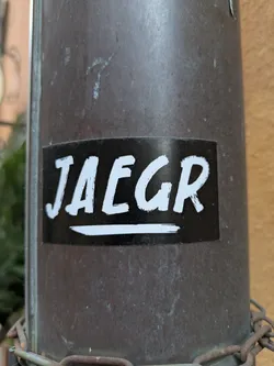 A black and white sticker with the word JAEGR written in a stylized, brush-stroke font. The sticker is applied to a dark brown, cylindrical metal surface.